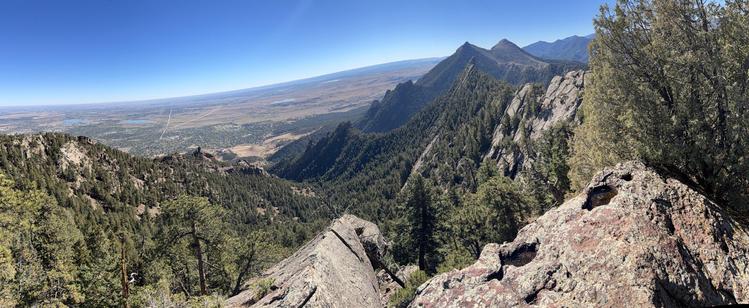 A panoramic view of a mountainous landscape featuring rugged peaks, lush greenery, and a clear blue sky. The foreground includes rocky terrain with scattered trees, while the background showcases a vast valley and distant mountains.