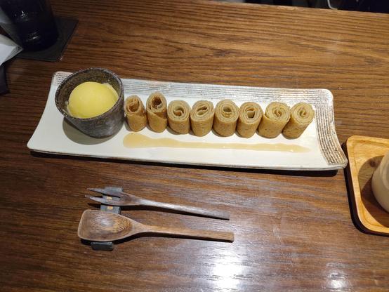 Photo d'une assiette rectangulaire sur laquelle est posée une crêpe roulée et coupée en 8, avec une boule de sorbet yuzu et un trait de miel de pomme.