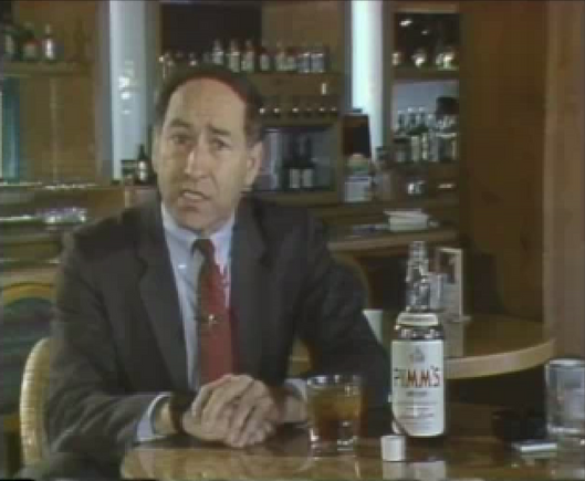 Stewart Cheifet sitting at a bar with a bottle of PIMS English gin on the table, along with some of the gin poured into a glass.