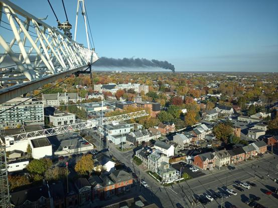 Distant view of Kingston, Ontario on a sunny, fall day, with a crane jib in the left foreground and a black column of smoke in the background the is levelled off at a height as it is blown south.