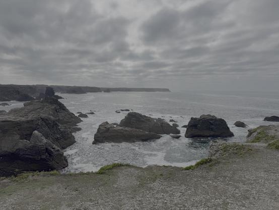 Dans une île Belle, une côte rocheuse très déchiquetée sur l'Océan à marée haute. Du promontoire où se trouve la photographe on voit des énormes rochers schisteux noirs dans l'eau et au fond une avancée rocheuse couverte de lande. Il fait gris, le ciel est chargé de nuages.