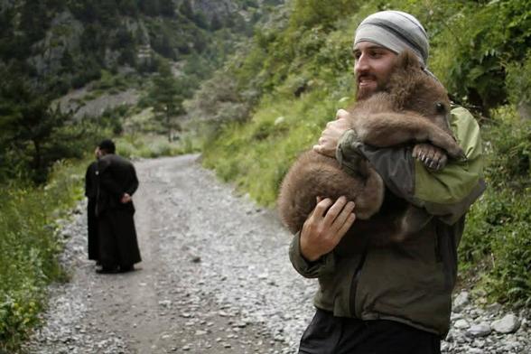A man cuddles a baby bear. In the background, an orthodox priest looms.