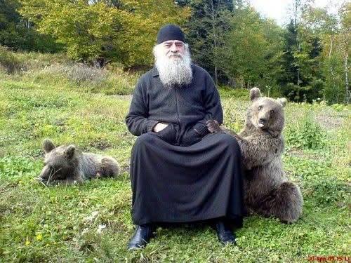 An orthodox priest sits in a field. A bear has a paw on his lap. Another bear lies in the background.