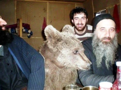 A tired-looking orthodox priest sits at a bar. Next to him is an equally tired-looking bear. Behind them, a grinning man photobombs