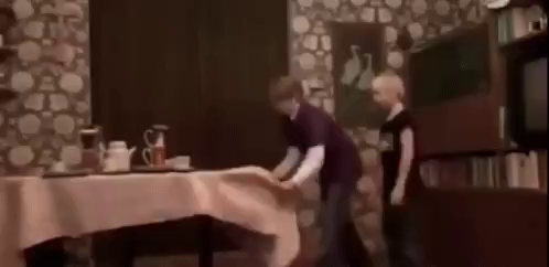 two youngsters pull a tablecloth from under some dishes, succeed, but one of them hits a cupboard which topples