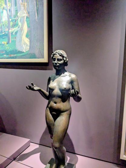A bronze statue of a nude woman standing against a muted purple wall in a museum gallery. Her head is crowned with a wreath of leaves or flowers, and her hands are raised slightly forward with open palms facing upward. To her left, framed on the wall, hangs a painting depicting figures in a wooded outdoor setting, including a central woman in a light yellow dress holding a tennis racket.