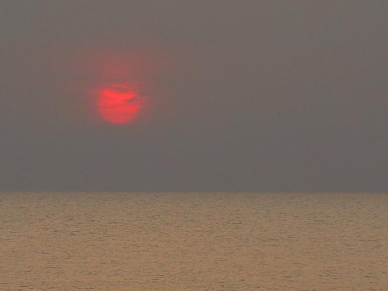 A tranquil seascape shows the sun emerging above the horizon over the Gulf of Thailand. The sun appears as a vivid red-orange disc softened by haze and partially veiled by thin cloud bands. The sky is muted grey with a misty atmosphere that diffuses the sunlight. Below, the calm sea occupies the lower third of the image, its surface lightly textured and tinted with warm golden-grey tones. The minimalist composition emphasises the contrast between the glowing sun and the subdued colours of the surrounding sky and water.
