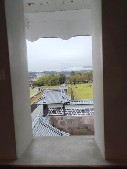 Vue d'une partie des toits du château typiquement japonais, au travers d'une fenêtre sans vitre. Au loin, on voit le parc du château et des arbres.