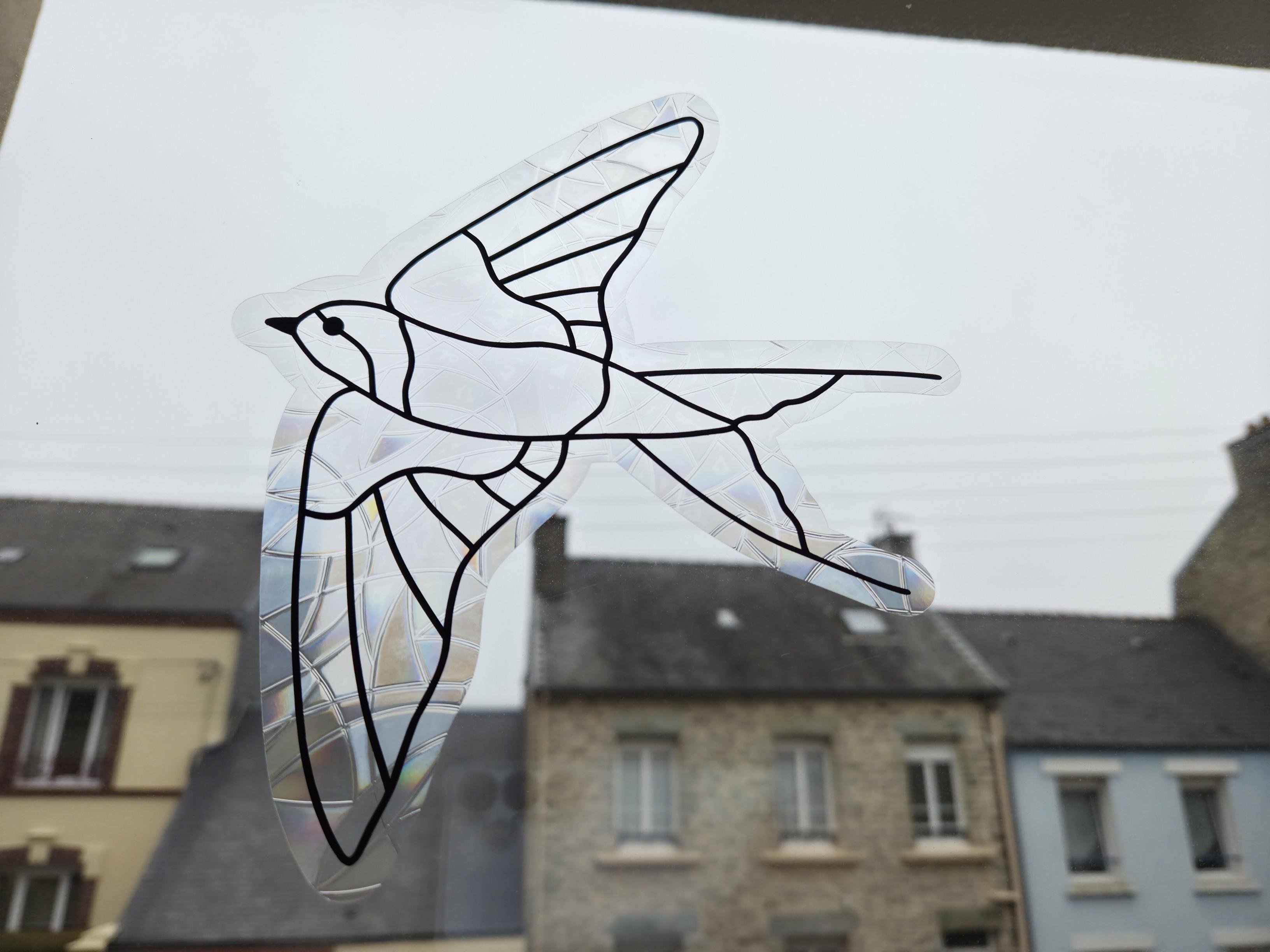 Un joli sticker en forme d'oiseau est collé sur une vitre. On voit le ciel et les toits des maisons d'en face.