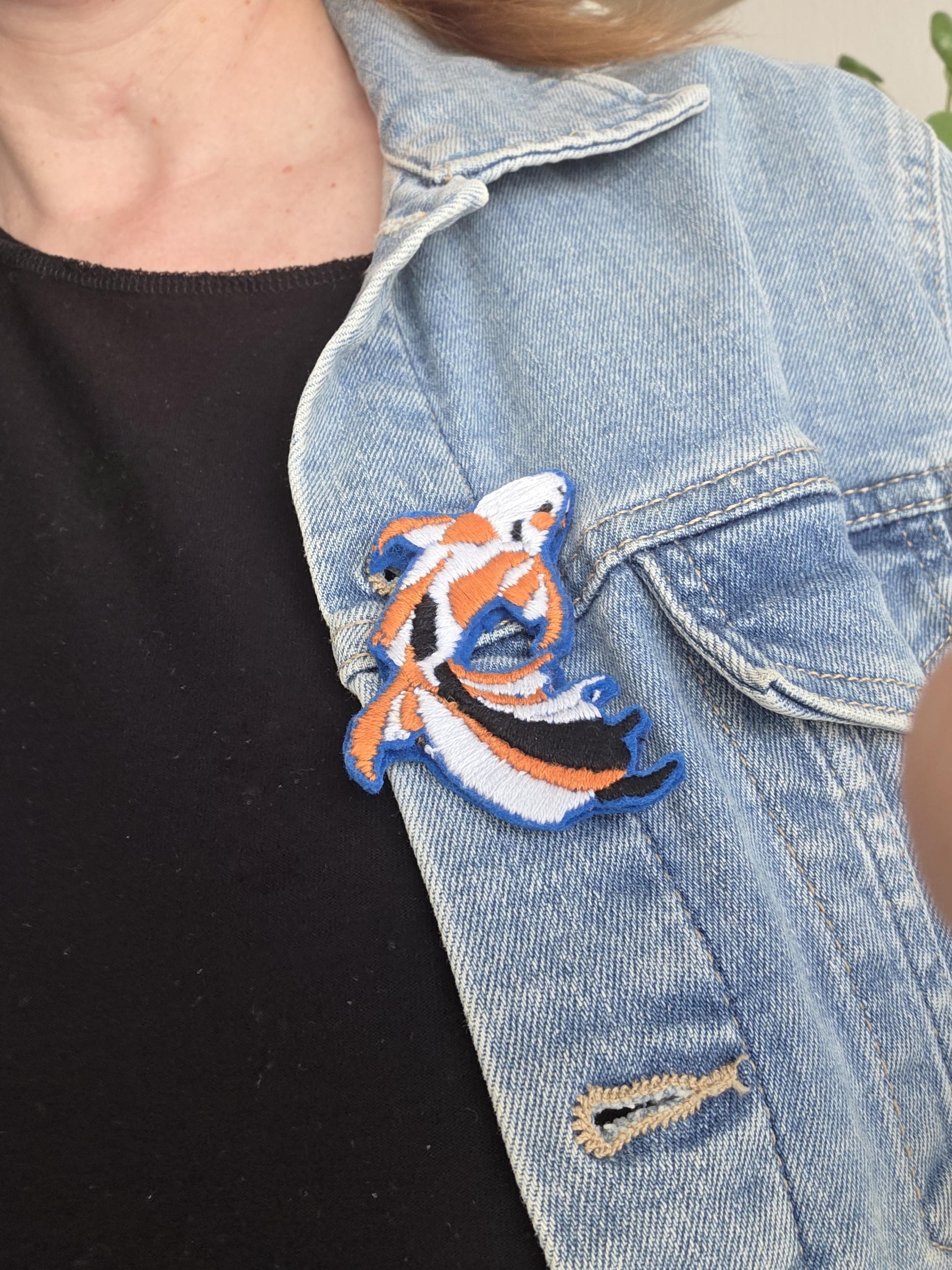 Broche en forme de poisson orange, blanc et noir. Elle est accrochée sur une veste en jean bleu clair.